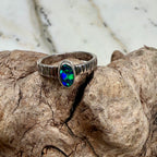 Aurora Reef Australian boulder opal ring on weathered driftwood beside the ocean, showing turquoise, green and deep blue flashes.