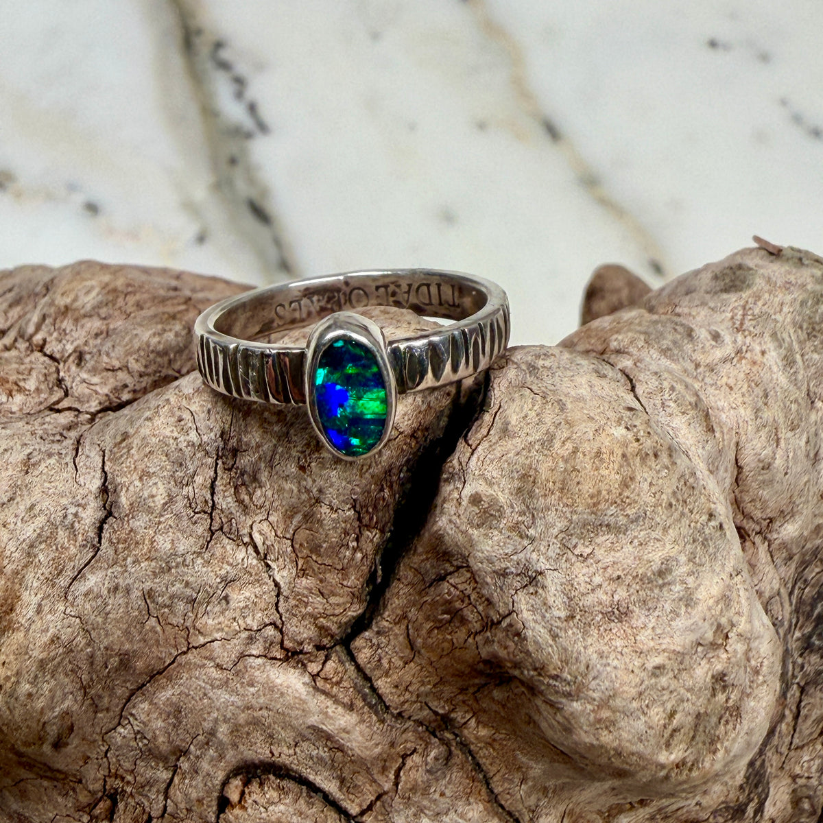 Aurora Reef Australian boulder opal ring on weathered driftwood beside the ocean, showing turquoise, green and deep blue flashes.