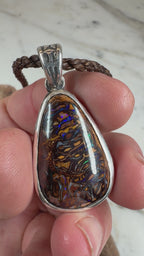 Ocean Relic Koroit Boulder Opal pendant with engraved sterling silver bezel displayed on rippled beach sand beside the ocean.